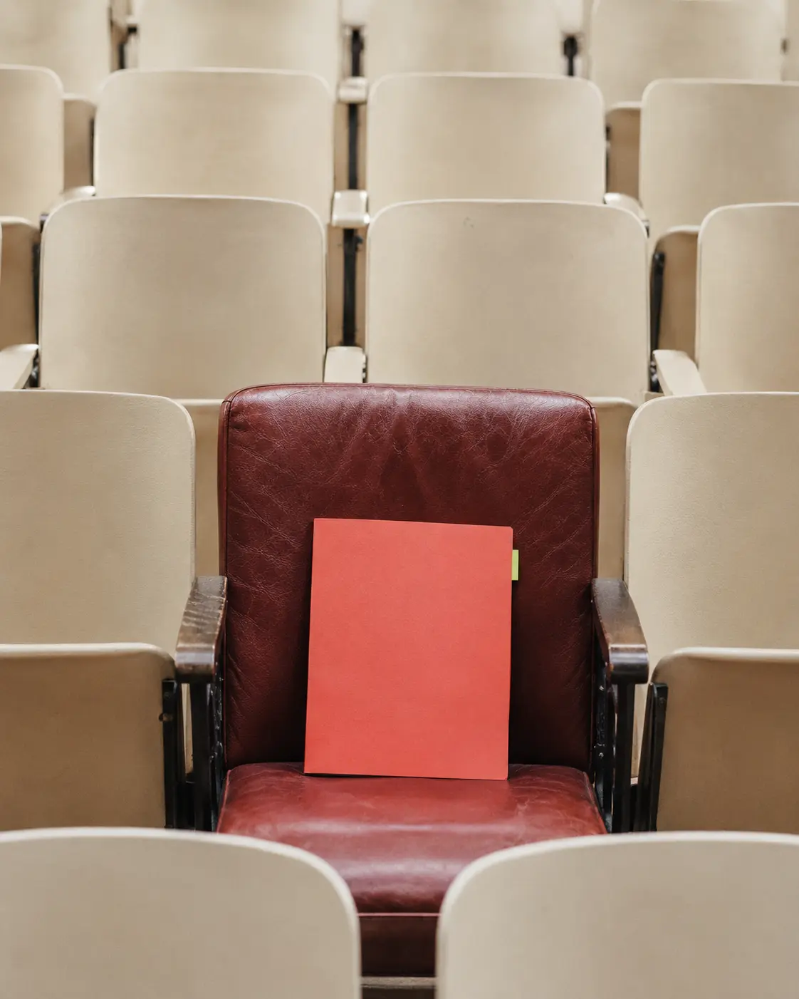 A coral project folder placed on a single oxblood-red seat in an ivory auditorium